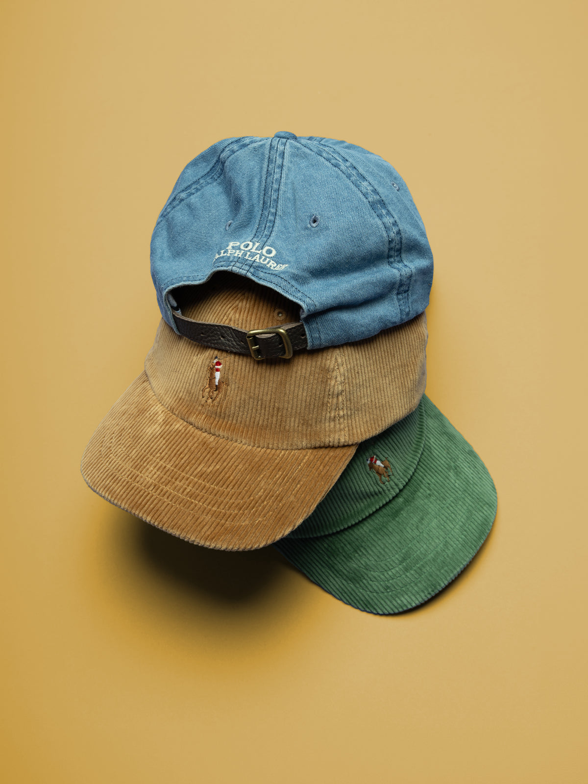 Polo Ralph Lauren corduroy caps in blue, tan and green with embroidered Polo logos, stacked against a mustard background